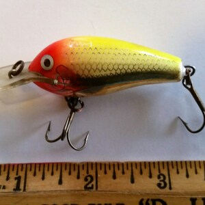 May include: A colorful fishing lure with a red head, yellow body, and silver scales. It has a clear plastic lip and two treble hooks. A wooden ruler with inch markings is in the foreground.