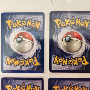 May include: Several Pok&eacute;mon trading cards are displayed, showcasing the reverse side design. Each card features the Pok&eacute;mon logo in yellow, a red and white Pok&eacute; Ball, and swirling blue and white graphics. A ruler is visible on the left side, with measurements in inches.