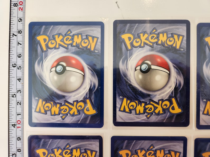 May include: Several Pok&eacute;mon trading cards are displayed, showcasing the reverse side design. Each card features the Pok&eacute;mon logo in yellow, a red and white Pok&eacute; Ball, and swirling blue and white graphics. A ruler is visible on the left side, with measurements in inches.