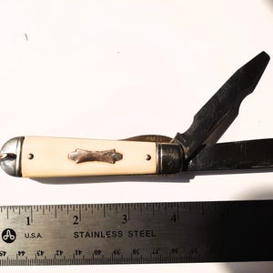 May include: A vintage pocket knife with a cream-colored handle and a decorative silver-toned inlay. The knife has two open blades and a small ring at the end. A stainless steel ruler is placed next to the knife.