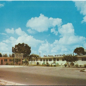 Könnte beinhalten: Eine Vintage-Postkartenansicht des Oak Crest Motels, einem einstöckigen rosa Gebäude mit Flachdach. Ein Schild im Vordergrund zeigt die Aufschrift Rooms Overnight, Kitchens, Low Rates. Die Szene liegt unter einem hellblauen Himmel mit großen, weißen Wolken.