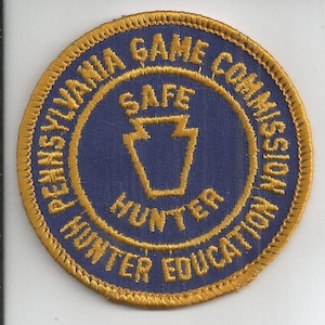 May include: A circular embroidered patch with a blue background and a gold border. The text reads Pennsylvania Game Commission Hunter Education in a circle, with the words Safe Hunter and a keystone outline in the center in gold thread.
