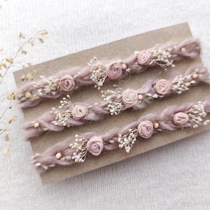 Può includere: Tre fasce per capelli intrecciate di colore malva chiaro, decorate con piccole rose di stoffa, delicati rametti bianchi e minuscole perline rosa. Le fasce sono esposte su un cartoncino dai toni neutri.