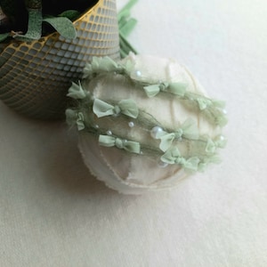Op de afbeelding: Een zacht, gebroken wit rekwisiet voor pasgeboren fotografie met delicate saliegroene lint- en strikaccenten. Kleine imitatieparels zijn langs het lint verspreid. Het rekwisiet wordt waarschijnlijk gebruikt voor fotoshoots van baby's.