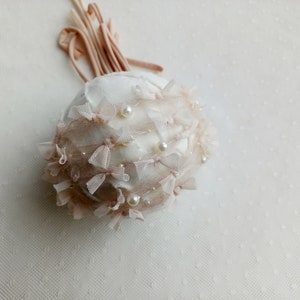 Puede incluir: Adorno decorativo redondo blanco roto adornado con peque&ntilde;os lazos rosas, perlas y delicado encaje. Un manojo de cintas rosas a juego se extiende desde la parte superior. El adorno tiene un aspecto suave y texturizado.