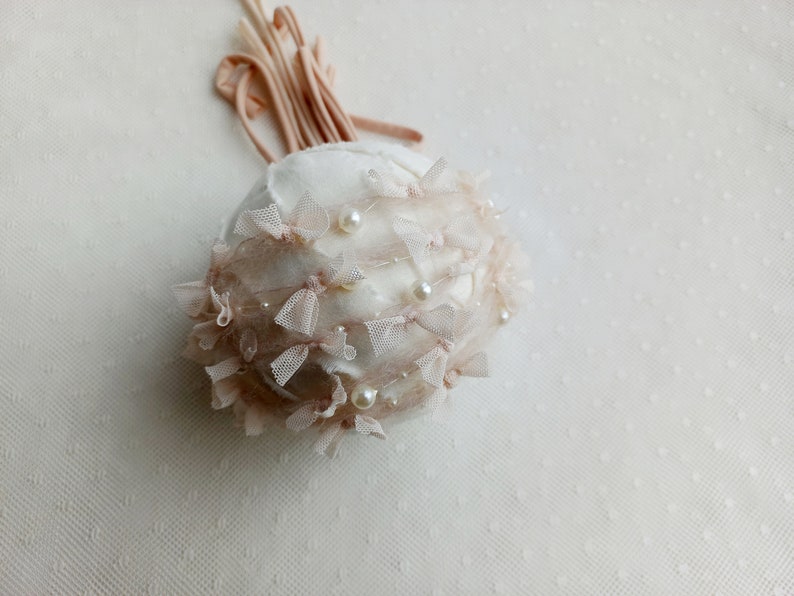 Puede incluir: Adorno decorativo redondo blanco roto adornado con peque&ntilde;os lazos rosas, perlas y delicado encaje. Un manojo de cintas rosas a juego se extiende desde la parte superior. El adorno tiene un aspecto suave y texturizado.