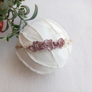 May include: A delicate headband with a row of mauve fabric flowers and gold beads. The headband is on a white fabric background.