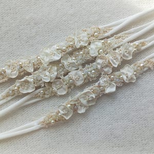 May include: Several white headbands with floral and pearl embellishments. Each headband features small, delicate fabric flowers, seed beads, and faux pearls. The headbands are arranged diagonally against a textured, off-white background.