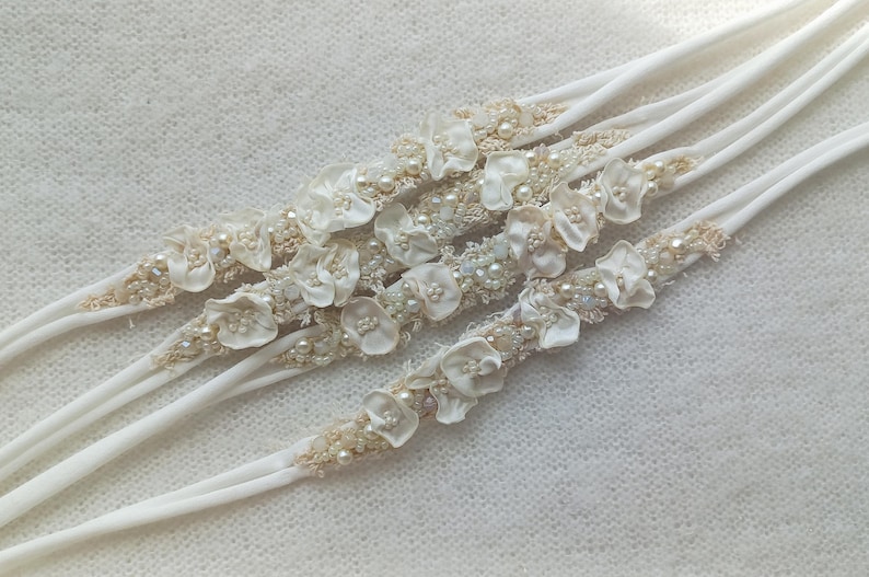May include: Several white headbands with floral and pearl embellishments. Each headband features small, delicate fabric flowers, seed beads, and faux pearls. The headbands are arranged diagonally against a textured, off-white background.