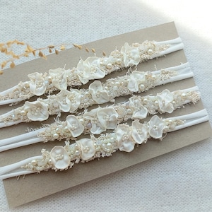 May include: Four ivory-colored headbands with floral and pearl embellishments. Each headband features a white elastic band and a decorative section with fabric flowers, beads, and pearls. The headbands are arranged on a neutral-toned surface.