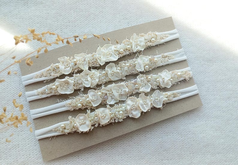 May include: Four ivory-colored headbands with floral and pearl embellishments. Each headband features a white elastic band and a decorative section with fabric flowers, beads, and pearls. The headbands are arranged on a neutral-toned surface.