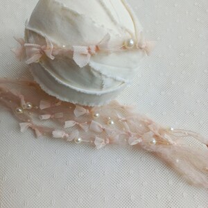 Puede incluir: Un accesorio de fotograf&iacute;a para reci&eacute;n nacidos de color crema, con una base redonda cubierta de tela y un borde deshilachado. Delicados detalles de encaje rosa y perlas adornan el accesorio, creando una est&eacute;tica suave y elegante. Una hebra de encaje rosa a juego con perlas se extiende desde la base.