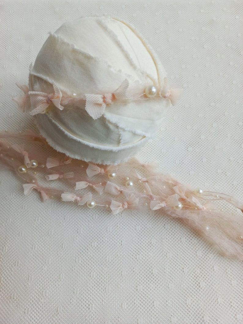 Puede incluir: Un accesorio de fotograf&iacute;a para reci&eacute;n nacidos de color crema, con una base redonda cubierta de tela y un borde deshilachado. Delicados detalles de encaje rosa y perlas adornan el accesorio, creando una est&eacute;tica suave y elegante. Una hebra de encaje rosa a juego con perlas se extiende desde la base.