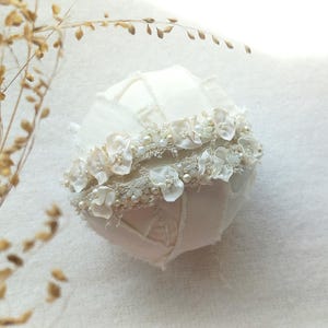 May include: A white, fabric-covered decorative sphere, adorned with delicate white floral embellishments and small pearls. The sphere has a patchwork design and is accented with a floral band. Dried, light brown foliage is in the background.
