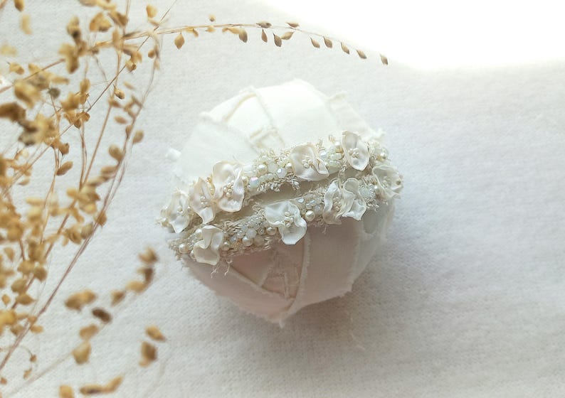 May include: A white, fabric-covered decorative sphere, adorned with delicate white floral embellishments and small pearls. The sphere has a patchwork design and is accented with a floral band. Dried, light brown foliage is in the background.