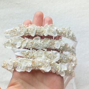 May include: A stack of ivory-colored headbands, each adorned with delicate fabric flowers and small, pearl-like beads. The headbands are held in a hand, showcasing their intricate details and soft, romantic aesthetic. Ideal for weddings or special occasions.