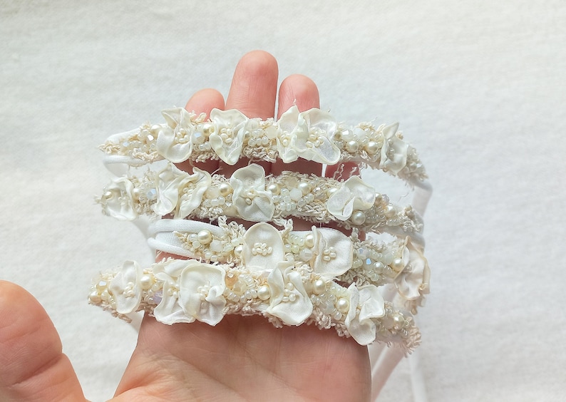 May include: A stack of ivory-colored headbands, each adorned with delicate fabric flowers and small, pearl-like beads. The headbands are held in a hand, showcasing their intricate details and soft, romantic aesthetic. Ideal for weddings or special occasions.