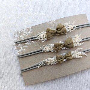 May include: Three baby headbands with olive green bows and dried floral accents. The headbands are displayed on a brown card, with a soft white background. The bows are made of a textured fabric.