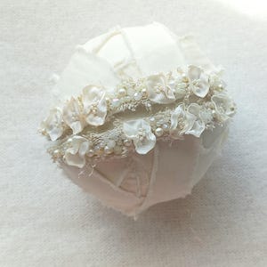 May include: A white, round decorative object, possibly a wedding accessory, with a textured surface. It is adorned with a band of small white flowers, pearls, and beads. The overall design is delicate and feminine.