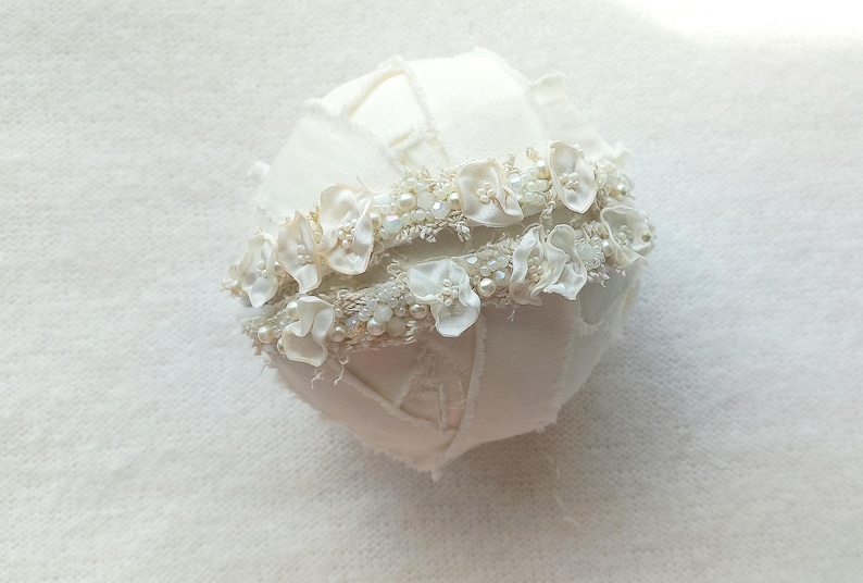 May include: A white, round decorative object, possibly a wedding accessory, with a textured surface. It is adorned with a band of small white flowers, pearls, and beads. The overall design is delicate and feminine.
