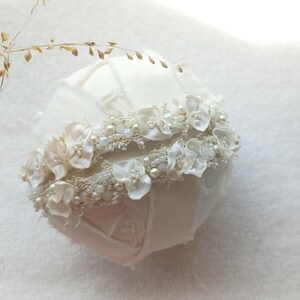 May include: A white, fabric-covered sphere, adorned with a band of delicate white flowers, pearls, and beads. The sphere has a patchwork design. Dried, brown sprigs are in the upper left corner. The overall theme is bridal or wedding decor.