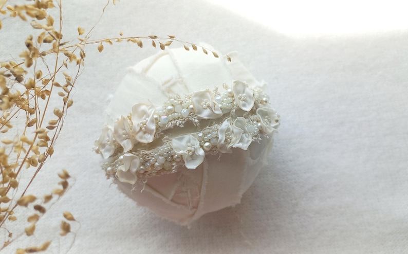 May include: A white, fabric-covered sphere, adorned with a band of delicate white flowers, pearls, and beads. The sphere has a patchwork design. Dried, brown sprigs are in the upper left corner. The overall theme is bridal or wedding decor.