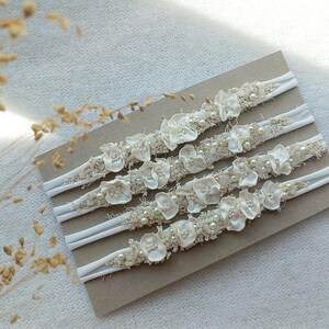May include: Five ivory-colored headbands with floral and pearl embellishments are displayed on a brown card. The headbands have a white elastic band and are decorated with small, delicate flowers and faux pearls.