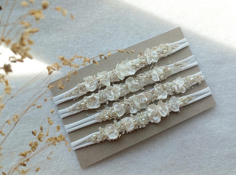 May include: Five ivory-colored headbands with floral and pearl embellishments are displayed on a brown card. The headbands have a white elastic band and are decorated with small, delicate flowers and faux pearls.