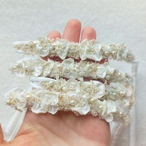 May include: Several white headbands with floral and pearl embellishments are held in a hand. The headbands feature fabric flowers, small pearls, and beaded accents. The headbands are likely for a special occasion.