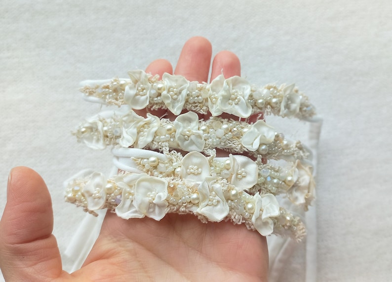May include: Several white headbands with floral and pearl embellishments are held in a hand. The headbands feature fabric flowers, small pearls, and beaded accents. The headbands are likely for a special occasion.