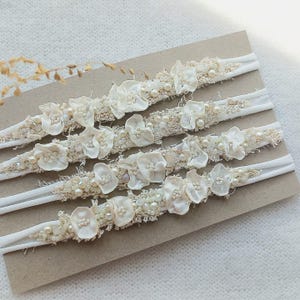 May include: Five ivory-colored headbands with floral and pearl embellishments. Each headband features delicate fabric flowers and small pearls, arranged on a textured band. The headbands are displayed on a neutral-toned surface.