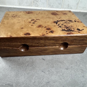 K&ouml;nnte beinhalten: Eine Holzkiste mit einem Furnierholzdeckel und einem dunklen Holzsockel. Der Deckel hat ein eingebranntes Fischdesign. Die Kiste hat zwei kleine runde L&ouml;cher an der Vorderseite.