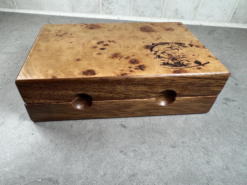K&ouml;nnte beinhalten: Eine Holzkiste mit einem Furnierholzdeckel und einem dunklen Holzsockel. Der Deckel hat ein eingebranntes Fischdesign. Die Kiste hat zwei kleine runde L&ouml;cher an der Vorderseite.