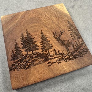 May include: A square wooden chopping board with an engraved forest scene. The design features pine trees, rocks, and an elk with antlers. The wood has a warm, natural brown tone.