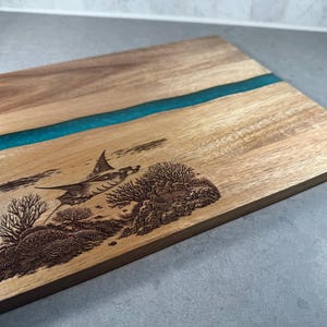 Puede incluir: Tabla de cortar de madera con un diseño de río de resina azul turquesa. La tabla presenta una ilustración grabada de una manta raya nadando sobre corales. La madera tiene una veta natural y una forma rectangular.