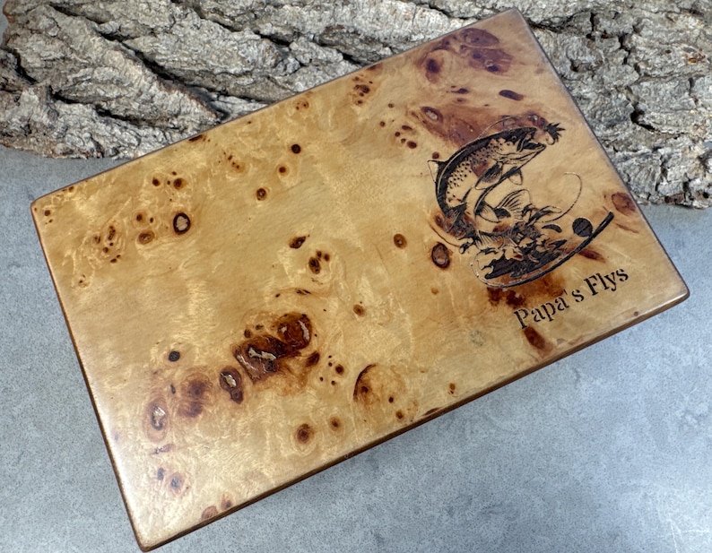 K&ouml;nnte beinhalten: Eine Holzkiste mit einer nat&uuml;rlichen Holzmaserung. Die Kiste hat ein eingebranntes Bild eines springenden Fisches und den Text "Papa's Flys".