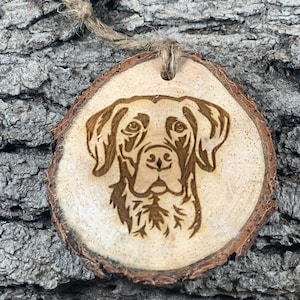 May include: A wooden ornament featuring a laser-engraved image of a dog's head. The ornament is made from a natural wood slice and has a rustic, handcrafted look.