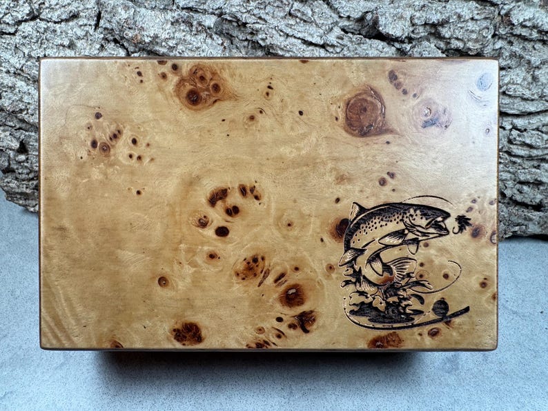 K&ouml;nnte beinhalten: Eine Holzkiste mit einer hellbraunen Maserung. Die Kiste hat ein eingebranntes schwarzes Design eines Fisches, der mit einer Fliege im Maul aus dem Wasser springt.
