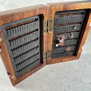 K&ouml;nnte beinhalten: Eine h&ouml;lzerne Fliegenfischbox mit einem Scharnierdeckel, ge&ouml;ffnet, um Reihen von Schlitzen zur Aufbewahrung von Fliegen zu zeigen. Die Box ist aus dunklem Holz mit einer nat&uuml;rlichen Oberfl&auml;che gefertigt. Mehrere Fliegen sind in den Schlitzen sichtbar.