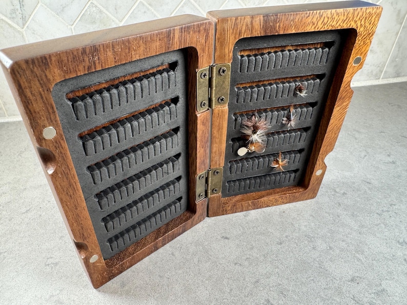 K&ouml;nnte beinhalten: Eine h&ouml;lzerne Fliegenfischbox mit einem Scharnierdeckel, ge&ouml;ffnet, um Reihen von Schlitzen zur Aufbewahrung von Fliegen zu zeigen. Die Box ist aus dunklem Holz mit einer nat&uuml;rlichen Oberfl&auml;che gefertigt. Mehrere Fliegen sind in den Schlitzen sichtbar.
