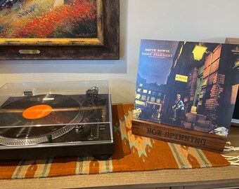 May include: A black turntable with a clear cover and a record playing. Next to it is a wooden stand holding a vinyl record album titled "Ziggy Stardust" by David Bowie. The stand reads "Now Spinning".