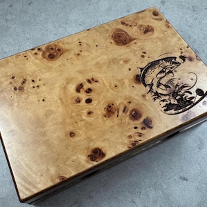 K&ouml;nnte beinhalten: Eine Holzkiste mit dunkelbrauner Oberfl&auml;che und einem hellbraunen Holzmaserungsmuster. Die Oberseite der Kiste ist mit einem Laser-gravierten Bild eines springenden Fisches verziert.
