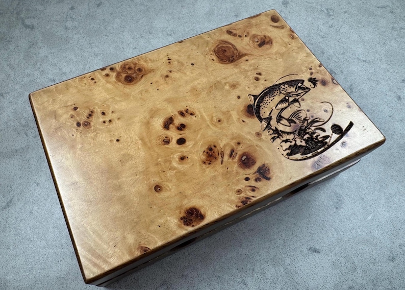 K&ouml;nnte beinhalten: Eine Holzkiste mit dunkelbrauner Oberfl&auml;che und einem hellbraunen Holzmaserungsmuster. Die Oberseite der Kiste ist mit einem Laser-gravierten Bild eines springenden Fisches verziert.