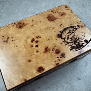 K&ouml;nnte beinhalten: Eine Holzkiste mit einer Maserung aus Wurzelholz und einem Laser-gravierten Bild eines springenden Fisches. Die Kiste hat einen Klappdeckel und ein dunkelbraunes Innenleben.