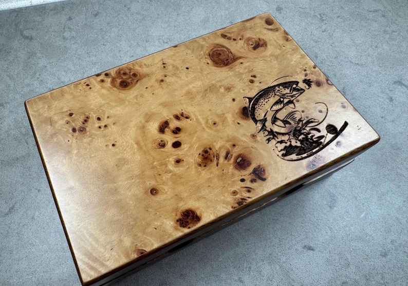 K&ouml;nnte beinhalten: Eine Holzkiste mit einer Maserung aus Wurzelholz und einem Laser-gravierten Bild eines springenden Fisches. Die Kiste hat einen Klappdeckel und ein dunkelbraunes Innenleben.