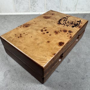 K&ouml;nnte beinhalten: Eine Holzkiste mit einer hellbraunen Maserholz-Oberseite und einem dunkelbraunen Holzsockel. Die Oberseite ist mit einem gravierten Design eines springenden Fisches verziert. Die Kiste hat zwei kleine L&ouml;cher an der Seite.