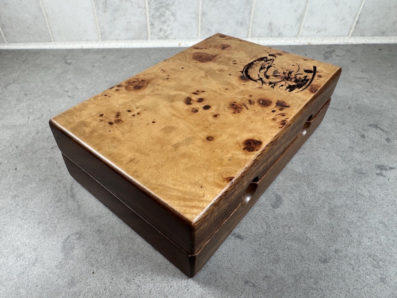 K&ouml;nnte beinhalten: Eine Holzkiste mit einer hellbraunen Maserholz-Oberseite und einem dunkelbraunen Holzsockel. Die Oberseite ist mit einem gravierten Design eines springenden Fisches verziert. Die Kiste hat zwei kleine L&ouml;cher an der Seite.