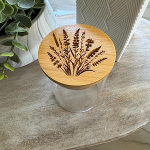 May include: A clear glass jar with a wooden lid featuring an engraved botanical design. The lid is round and made of light-coloured wood. The jar is sitting on a light-coloured wooden surface.