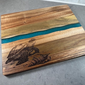 Puede incluir: Tabla de cortar de madera con un diseño de tortuga marina y concha grabado con láser. Un río de resina turquesa atraviesa el centro, añadiendo un toque de color. La tabla es rectangular y está hecha de madera clara y oscura.