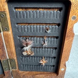K&ouml;nnte beinhalten: Eine h&ouml;lzerne Fliegenfischbox mit einem schwarzen Schaumstoffeinsatz. Die Box ist ge&ouml;ffnet und es befinden sich mehrere Fliegen im Schaumstoff.
